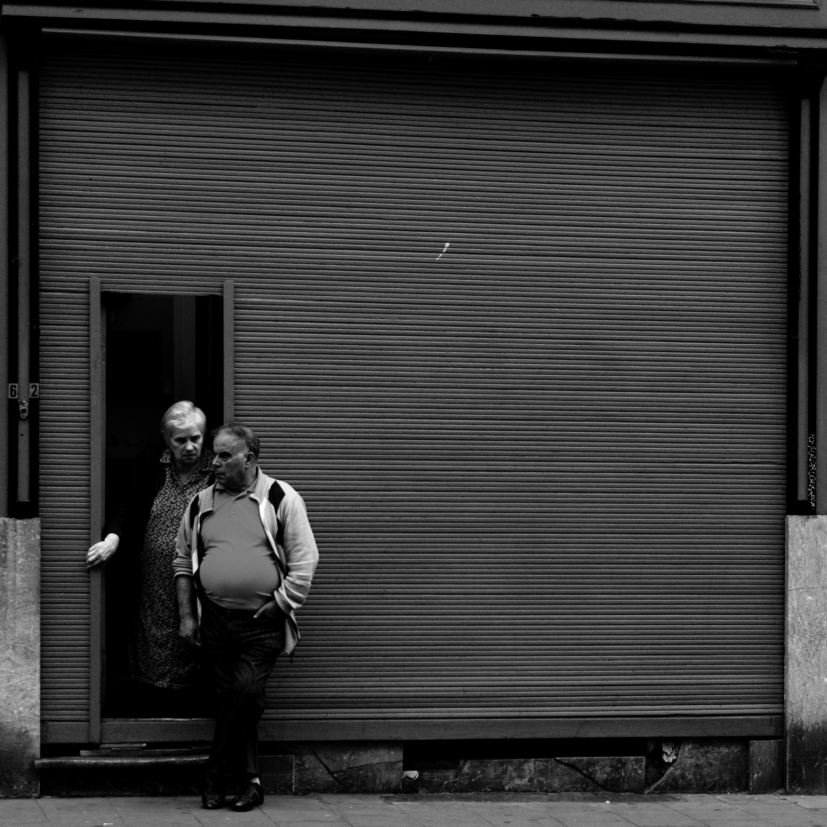 Older couple beside a dark shuttered doorway in black and white.